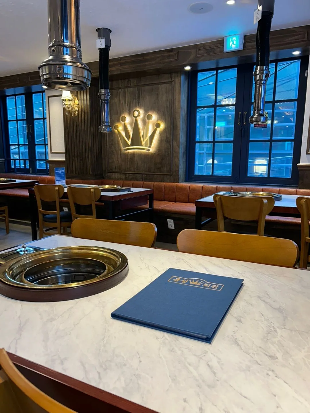 Illuminated gold crown emblem on the hall wall, with a brass grill plate and a navy gilded menu cover on a white marble table